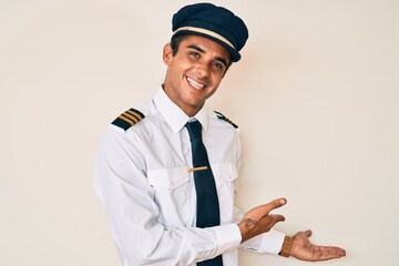 Young hispanic man wearing airplane pilot uniform inviting to enter smiling natural with open hand