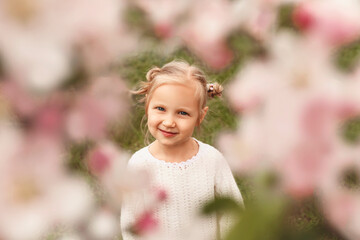 Blonde little girl photographed through the flowering branches of a tree. Child in the spring flower garden