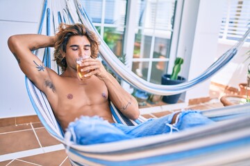 Young hispanic man relaxed drinking beer lying on the hammock at terrace.