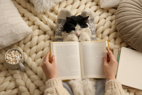 Woman reading book and holding adorable cat on knitted blanket, top view