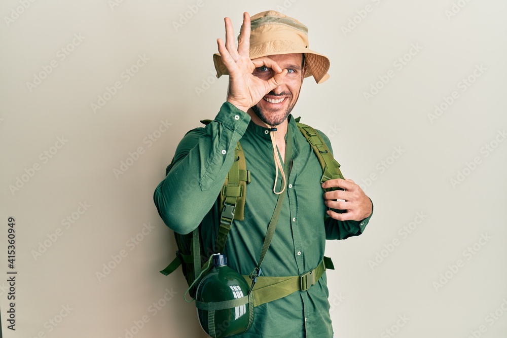 Wall mural Handsome man with beard wearing explorer hat and backpack doing ok gesture with hand smiling, eye looking through fingers with happy face.