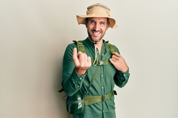 Handsome man with beard wearing explorer hat and backpack beckoning come here gesture with hand inviting welcoming happy and smiling