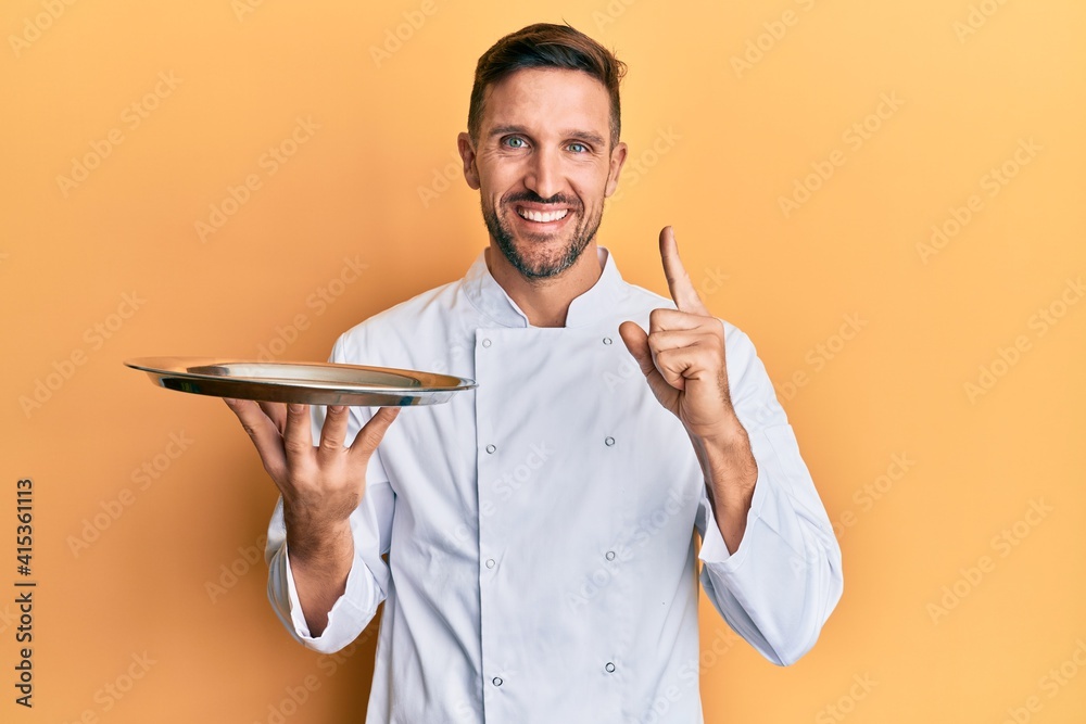 Wall mural Handsome man with beard wearing chef uniform holding silver tray smiling with an idea or question pointing finger with happy face, number one