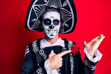 Young man wearing mexican day of the dead costume over red amazed and smiling to the camera while presenting with hand and pointing with finger.