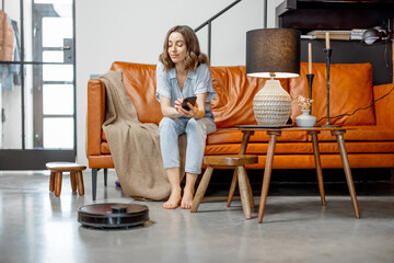 Black robotic vacuum cleaner cleaning the floor while woman sitting on sofa and looking on her...