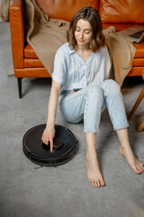 Black robotic vacuum cleaner cleaning the floor while woman sitting near sofa and relax. Smart...
