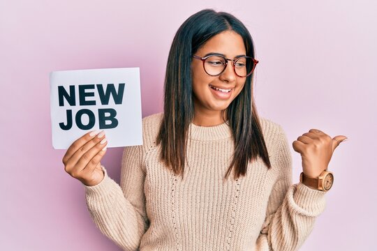 Young latin girl holding new job message on paper pointing thumb up to the side smiling happy with open mouth
