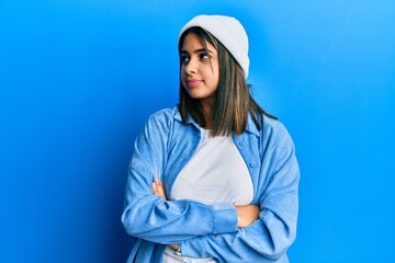 Young latin woman wearing cute wool cap looking to the side with arms crossed convinced and...