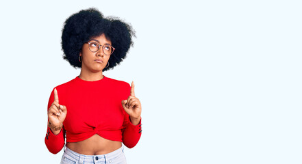 Young african american girl wearing casual clothes and glasses pointing up looking sad and upset, indicating direction with fingers, unhappy and depressed.