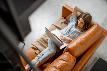 Young woman working with laptop while lying on foxy leather sofa. Digital nomad and work from home...