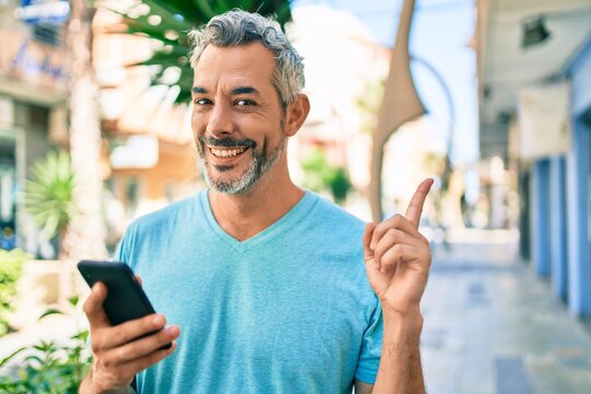 Middle age grey-haired man using smartphone at street of city smiling happy pointing with hand and finger to the side