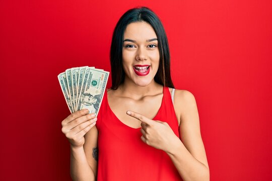 Young hispanic girl holding 20 dollars banknote smiling happy pointing with hand and finger