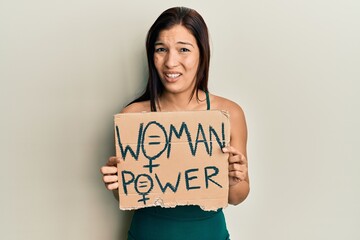 Young latin woman holding woman power banner clueless and confused expression. doubt concept.