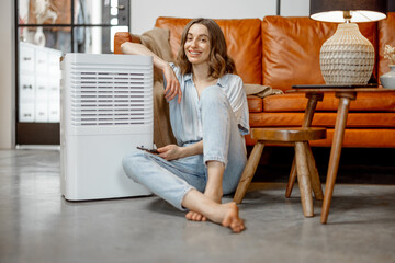 Pretty woman sitting near air purifier and moisturizer appliance near sofa monitoring air quality...