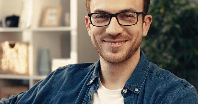 Close-up portrait of handsome bearded man with glasses sitting at home on sofa and looking at camera smiling.