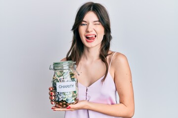 Young beautiful caucasian girl holding charity jar with money winking looking at the camera with...