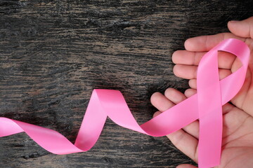 Top view of hand holding bright pink ribbon on dark wood background. Breast cancer awareness and...