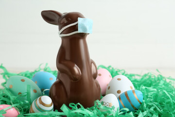 Chocolate bunny with protective mask and eggs against white background, closeup. Easter holiday during