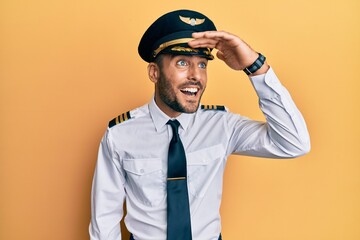 Handsome hispanic man wearing airplane pilot uniform very happy and smiling looking far away with hand over head. searching concept.