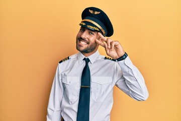 Handsome hispanic man wearing airplane pilot uniform doing peace symbol with fingers over face, smiling cheerful showing victory