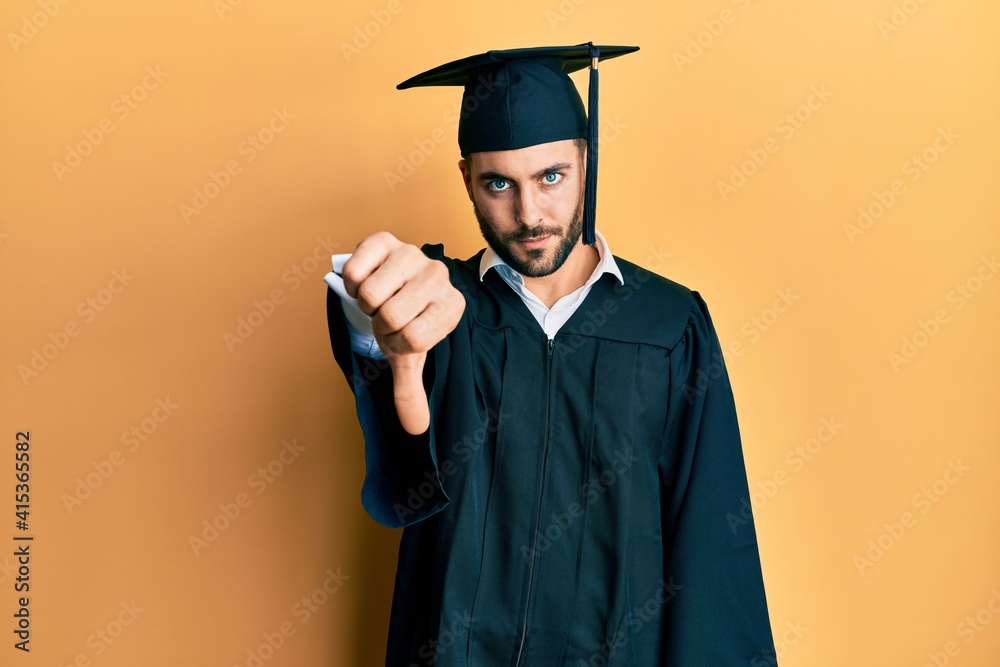 Sticker Young hispanic man wearing graduation cap and ceremony robe looking unhappy and angry showing rejection and negative with thumbs down gesture. bad expression.