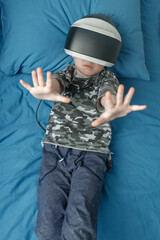 Five-year-old cute boy is lying in his bed in VR glasses and playing 3D games with his hands outstretched in front of him. concept of new technologies. Copy space