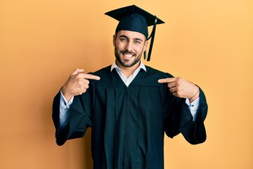 Young hispanic man wearing graduation cap and ceremony robe looking confident with smile on face, pointing oneself with fingers proud and happy.