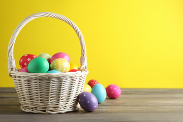 Colorful Easter eggs in wicker basket on wooden table against yellow background. Space for text