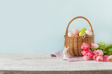 Easter wicker basket with painted eggs and pink tulips on blue background with space for text.