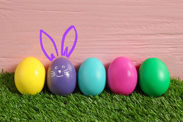 Violet eggs with drawn face and ears as Easter bunny among others on green grass against pink wooden background