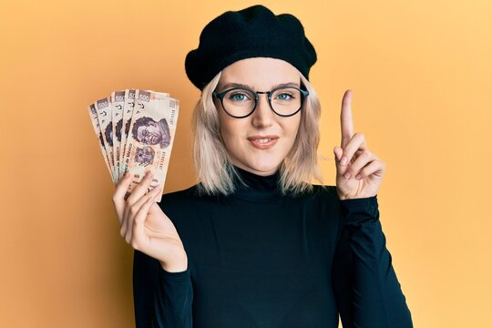 Young blonde girl holding mexican pesos smiling with an idea or question pointing finger with happy face, number one