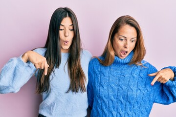 Hispanic family of mother and daughter wearing wool winter sweater pointing down with fingers showing advertisement, surprised face and open mouth