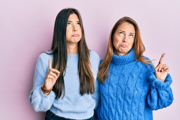 Hispanic family of mother and daughter wearing wool winter sweater pointing up looking sad and upset, indicating direction with fingers, unhappy and depressed.