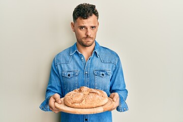 Young hispanic man holding bread skeptic and nervous, frowning upset because of problem. negative...