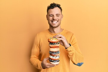 Young hispanic man holding tasty colorful doughnuts winking looking at the camera with sexy...