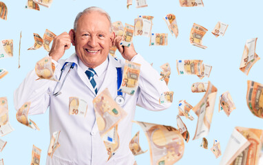 Senior handsome grey-haired man wearing doctor coat and stethoscope smiling pulling ears with...