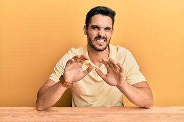 Young hispanic man wearing casual clothes sitting on the table disgusted expression, displeased and...