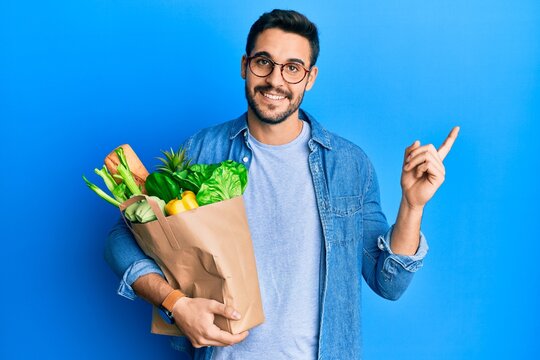 Young hispanic man holding paper bag with bread and groceries smiling happy pointing with hand and finger to the side