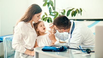 Fototapeta premium Male Doctor Pediatrician Examining an Ill Sad Kid Girl at Medical Visit With Mother in the Hospital. Male Family Doctor Examining and Consulting to Mother and Her Ill Child.