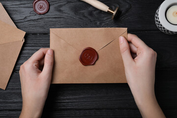 Woman holding envelope with wax seal at black wooden table, top view