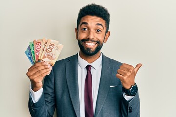 Handsome hispanic business man with beard holding canadian dollars pointing thumb up to the side smiling happy with open mouth