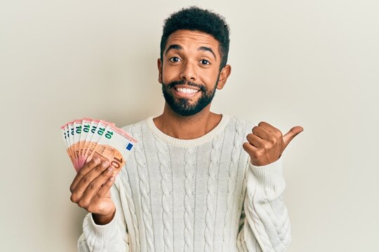 Handsome hispanic man with beard holding bunch of 10 euro banknotes pointing thumb up to the side smiling happy with open mouth