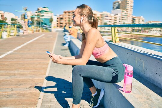 Young blonde sportswoman doing exercise using smartphone sitting on the bench at the promenade.