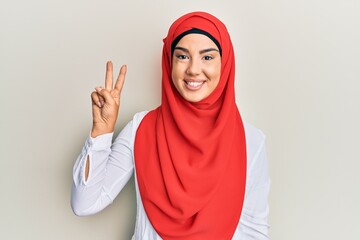 Young beautiful hispanic girl wearing traditional islamic hijab scarf showing and pointing up with fingers number two while smiling confident and happy.