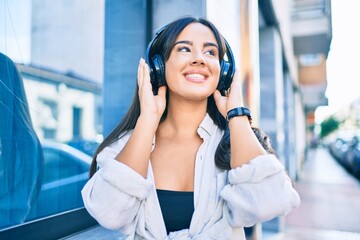 Young hispanic girl smiling happy listening to music using headphones at the city.