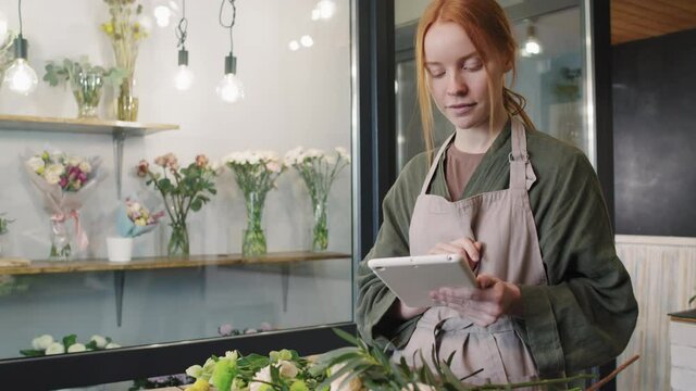 Tilting-up medium shot of pretty woman working in small flower shop doing flower inventory entering data on digital tablet