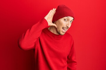 Bald man with beard wearing wool sweater and winter hat smiling with hand over ear listening and hearing to rumor or gossip. deafness concept.