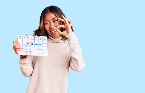 Young beautiful mixed race woman holding travel calendar doing ok sign with fingers, smiling friendly gesturing excellent symbol