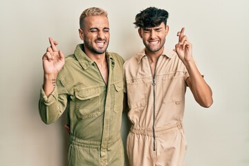 Homosexual gay couple standing together wearing casual jumpsuit gesturing finger crossed smiling with hope and eyes closed. luck and superstitious concept.