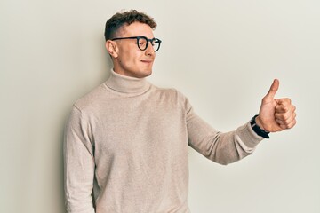 Hispanic young man wearing casual turtleneck sweater looking proud, smiling doing thumbs up gesture to the side